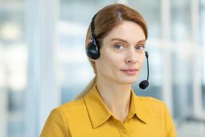 retrato de un joven mujer en un auriculares sentado en el oficina y mirando seriamente a el cámara. empleado de el línea directa, apoyo servicio. de cerca foto. foto