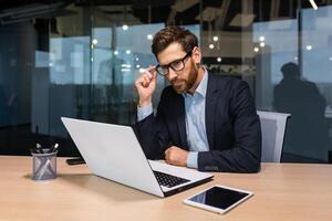 Serious thinking senior businessman working inside office using laptop, man in glasses solving problem, mature investor in business suit. photo