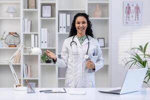 retrato de un joven hermosa clínica trabajador con un tableta computadora en su manos, el mujer sonrisas alegremente y mira a el cámara, soportes dentro el clínica oficina, invita para un cita y consulta. foto