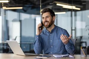 sonriente joven hombre empresario tiene un negocio conversacion con clientela y socios en el teléfono, se sienta en el oficina a el mesa y explica activamente gesticulando con su manos. foto