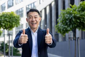 Smiling Asian businessman outdoors giving two thumbs up, expressing success and positivity in a corporate environment. photo