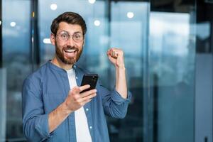 retrato de un moderno empresario en el medio de el oficina, el hombre es mirando a el cámara con un teléfono inteligente y celebrando el victoria, el hombre es participación el teléfono en su manos y levantamiento su mano en un gesto de triunfo. foto