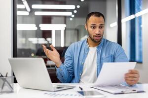 Surprised Indian man in a living room setting, dressed as a businessman, reacting to a paper document. Concept of unexpected news in home office environment. photo