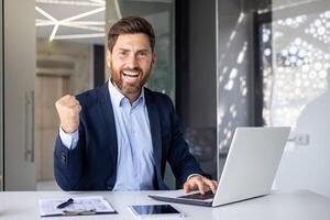 Portrait of a happy young businessman showing a hand gesture of success and victory to the camera. He sits in the office at the table and works on a laptop and with a project. photo