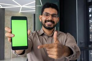 Happy indian male in eyeglasses directing with index finger on mobile phone screen with empty space. Young bearded man recommending new application and approving benefits of installing service. photo