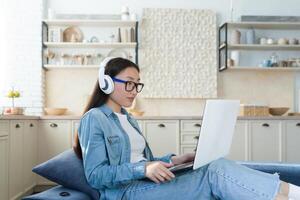 Young beautiful Asian teenage girl in headphones resting on sofa at home. Sitting on the couch, holding a laptop. He listens to music, watches a movie, spends the weekend photo