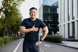 retrato de un asiático joven masculino deportista en pie en un ciudad calle y participación su cofre con su mano, sensación grave dolor, sobrecarga, mirando cansado a el cámara. foto