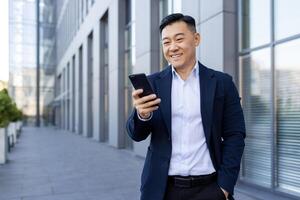 Smiling young asian business man lawyer standing outside office center and using mobile phone, hand in pocket. photo