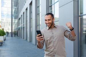 Happy african american businessman looking at smartphone screen, man in shirt outside modern office building received good news of win and success online, boss celebrating financial triumph photo