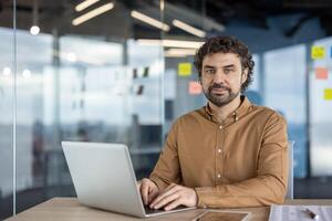 Portrait of a dedicated hispanic man using a laptop in a well-lit, contemporary office space, exuding professionalism and concentration. photo