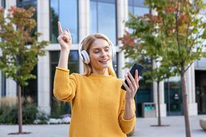 alegre rubia mujer en amarillo suéter bailes al aire libre mientras escuchando a música en su auriculares, utilizando un teléfono inteligente en un urbano ajuste. foto