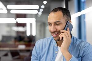 A cheerful man engages in a pleasant conversation on his mobile phone, with an office environment blurred in the background. photo