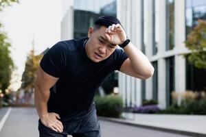 Serious young Asian male athlete and runner wiping sweat from forehead, suffering from headache, bent over from fatigue, resting. photo