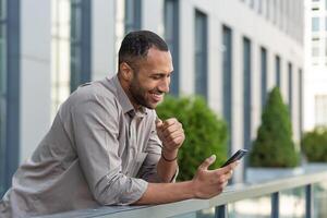 contento africano americano empresario mirando a teléfono inteligente pantalla, hombre en camisa fuera de moderno oficina edificio recibido bueno Noticias de ganar y éxito en línea, jefe celebrando financiero triunfo foto