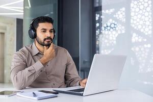 un grave pensando hindúes trabajos dentro un oficina con un computadora portátil, un hombre en auriculares escucha a podcasts y audio libros, un enfocado programador codifica nuevo software. foto