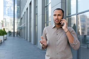 Outdoor portrait of angry and upset man outside office building, businessman in shirt talking on phone and gesturing with hands. photo