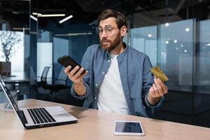 frustrado y engañado empresario en oficina molesto a hacer compra en en línea Tienda y en línea dinero transferir desde banco crédito tarjeta, irritado hombre trabajando dentro oficina utilizando ordenador portátil y teléfono. foto