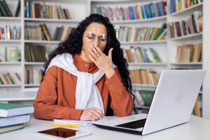 Sleepy and overtired young student studying in campus academic library until late, Hispanic woman preparing for entrance exams among books, using laptop to view online courses. photo