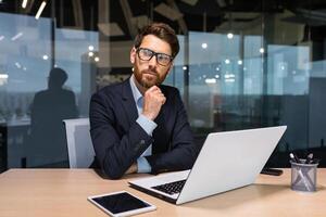 Serious thinking senior businessman working inside office using laptop, man in glasses solving problem, mature investor in business suit. photo