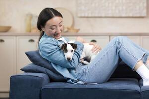 un sonriente joven hermosa asiático mujer es sentado en el sofá a hogar, participación un pequeño perro en su manos. él gasta su gratis hora con su mascota, jugando, descansando foto