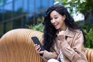 Young happy woman celebrating victory triumph success, satisfied hispanic business woman holding phone received online win notification, sitting on bench outside office building. photo