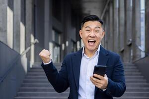 Portrait of a happy young Asian man in a business suit standing outside a building, holding a phone, looking at the camera and showing a victory gesture with his hand. photo