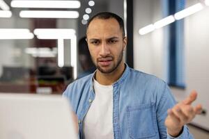 A young male professional in a blue denim shirt appears perplexed and frustrated while working at his computer in a modern office setting, expressing a common workplace challenge. photo