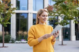 A professional businesswoman in a yellow sweater uses a digital tablet outside a modern office building. She looks content and focused. photo