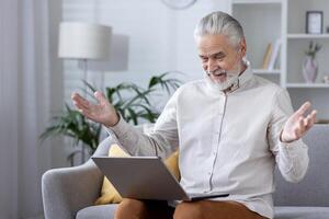 Elderly person sitting with open laptop gesturing a mix of surprise and confusion in a well-lit living room. photo