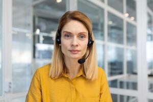 Web view camera, pov. Woman with a headset phone listens to the interlocutor, call, online meeting. The support service worker looks seriously and thoughtfully at camera, works inside office. photo