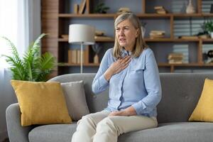 Senior woman touching chest while having pain in throat after coughing in domestic interior. Disturbed lady in white pants feel sick from virus or bacteria infection and having loss of voice. photo