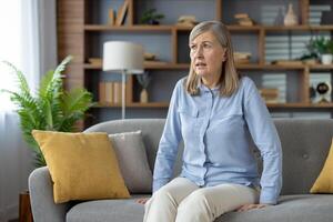 Stressed female in casual outfit sitting on grey sofa and looking aside with scared expression indoor. Frustrated woman experiencing anxiety attack or depression episode and mental health disorder. photo