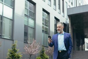 A senior man, a businessman in a suit, goes to a meeting on the street near a modern office center, holds a phone in his hand, looks at the phone, dials a number. He keeps his hand in his pocket. photo