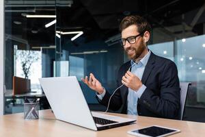 call successful businessman boss beard in business suit on online meeting, talking with colleagues using headset and laptop, for remote conversation, man working inside modern office building. photo