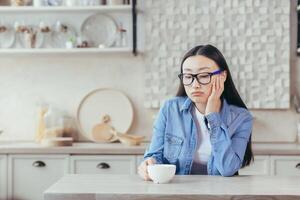 café descanso. joven cansado asiático mujer persona de libre dedicación. sentado a hogar a el cocina mesa con un taza de café, descansando, considerado. foto