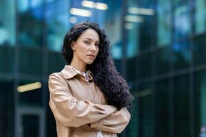 retrato de exitoso orgulloso mujer jefe fuera de oficina edificio, Hispano satisfecho con trabajo mirando a cámara con cruzado brazos, maduro mujer de negocios con Rizado pelo en pie y posando foto