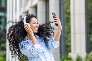 A young, carefree woman loses herself in the music, feeling the rhythm with large headphones and a smartphone in her hand, standing against the backdrop of an urban landscape. photo