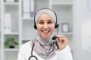 alegre hembra cuidado de la salud profesional vistiendo un hijab y auriculares en un llamar, atractivo con un paciente o colega, en un brillante oficina ajuste. foto