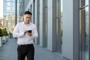 enfocado asiático empresario mensajes de texto en teléfono mientras caminando cerca vaso edificios urbano negocio estilo de vida concepto. foto