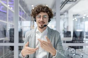 Curly-haired mature businessman with headset actively gestures while communicating in a brightly lit office environment. photo