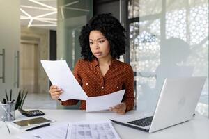 triste decepcionado mujer de negocios dentro oficina a lugar de trabajo mirando a documentos documentos financiero informes, africano americano hembra trabajador en papel trabajo Deprimido infeliz con financiero logros foto