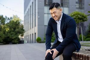 Contorted in pain, a young Asian businessman sits on a bench near an office center and holds his knee with his hands. Got an injury, inflammation. photo