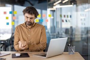 Male office worker experiencing discomfort, clutching his hand in pain at his workplace with laptop and stationary around. photo