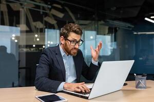 Angry mature boss got error laptop not turning on, program not working, senior man working on computer inside modern office building, businessman in glasses and business suit. photo