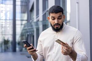 de cerca foto de un joven indio hombre en un camisa en pie afuera, participación un móvil teléfono y mirando confusamente a un crédito tarjeta