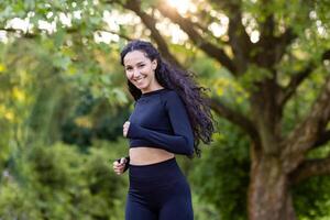 joven hermosa mujer corriendo en el parque, retrato de un latín americano mujer trotar y haciendo activo ejercicios, deportista sonriente y mirando a el cámara entre el arboles foto