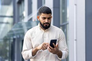 Photo of a serious Indian young man walking down the street near office buildings and using a mobile phone