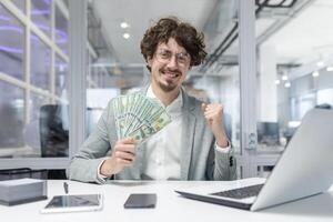 Cheerful young adult businessman holding cash at workplace, exuding confidence and financial success with a bright, satisfied smile. photo