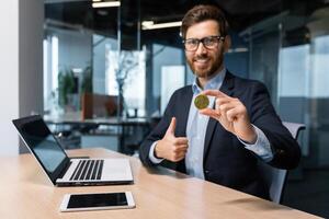 Portrait of mature successful businessman investor, man shows cryptocurrency gold coin to camera and thumbs up firmly, man works on stock exchange inside office online uses laptop. photo