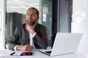 dolorido garganta frío, hombre dentro oficina trabajando con ordenador portátil a lugar de trabajo, empresario participación mano a garganta hizo una mueca en dolor. foto